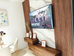 A modern living room with a large flat-screen TV mounted on a wooden wall, a glass coffee table, a white chair, and a small wooden side table with a framed certificate. - Luxury Stay in Maârif: High-Speed WiFi & Free Parking (Dar-el-Beida)