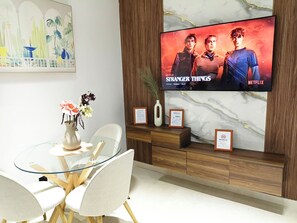 A modern living room with a flat-screen TV displaying 'Stranger Things,' a glass dining table, white chairs, a vase with flowers, and wooden cabinetry. - Luxury Stay in Maârif: High-Speed WiFi & Free Parking (Dar-el-Beida)
