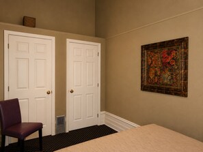 Ein Zimmer mit zwei weißen Türen, einem violetten Sessel, einem Bett mit gesteppter Bettdecke und einem gerahmten Kunstwerk an der Wand.