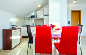 A modern kitchen with white cabinets, a microwave, and a coffee machine. There is a dining area with a table and red chairs. - (Podstrana)