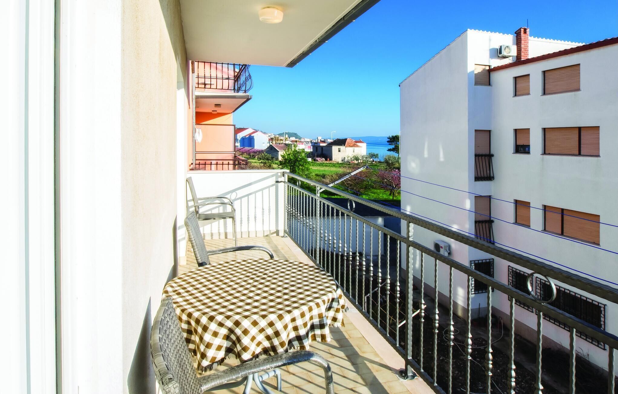 A balcony with a checkered table and chairs, overlooking a residential area and the sea.