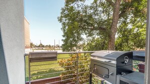 Ein Balkon mit Grill, Metallgeländer und Blick auf eine begrünte Fläche mit Bäumen.