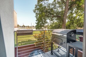 Ein Balkon mit Grill, Metallgeländer und Blick auf eine begrünte Fläche mit Bäumen.