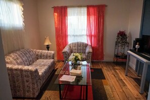 Ett vardagsrum med en mönstrad soffa, ett soffbord i glas med papper på, en lampa och en tv som står på ett TV-bord.