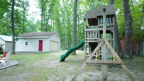 Eine hölzerne Spielkonstruktion mit Rutsche, einer roten Tür und einem kleinen Schuppen in einem Waldgebiet.