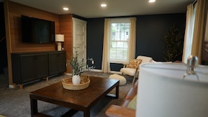 A living room with a dark wall, a television mounted above a wooden cabinet, a window with curtains, and a sofa with a cushion. - Big Blue at Little Long Lake (Harrison)