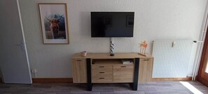 Ein Zimmer mit an der Wand befestigtem Fernseher, einem Holztisch mit Schubladen, einem gerahmten Bild an der Wand und einer Heizung.