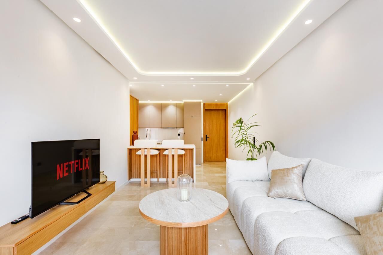 A modern living room with a flat-screen TV, a round coffee table, a sofa, and a kitchen area in the background.
