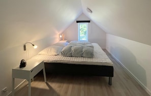 Quarto de sótão pequeno e bem iluminado, com uma cama de solteiro, mesa de cabeceira, escrivaninha e janela que permite entrada de luz natural.