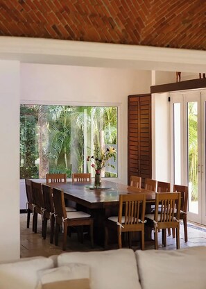 A dining area with a large table and chairs, a vase with flowers, and a view of tropical plants through a glass door. - Luxury Family Villa on Yalku Lagoon · Private Cenote · Akumal (Tulum)