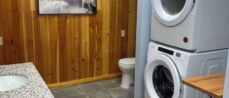 A bathroom with a wooden wall, a painting, a sink, a toilet, and stacked washing machines.