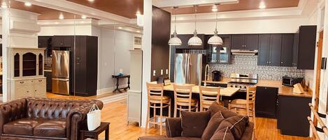 A spacious living area with a brown leather sofa, a coffee table, and a dining area with wooden chairs and a kitchen in the background.