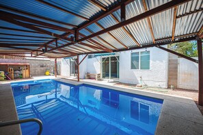 Zona de piscina techada con techo metálico y una casa al fondo.