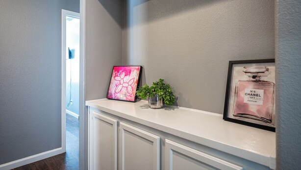 A hallway with a white console table displaying a pink abstract painting, a small potted plant, and a framed perfume bottle. - Cozy Cool 3 Bedrooms Home W~Pool (North Las Vegas)