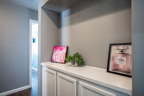 A hallway with a white console table displaying a pink abstract painting, a small potted plant, and a framed perfume bottle. - Cozy Cool 3 Bedrooms Home W~Pool (North Las Vegas)