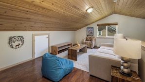 Una stanza in stile loft con soffitto spiovente, una panca di legno, un pouf blu, un divano bianco e una lampada su un tavolino.