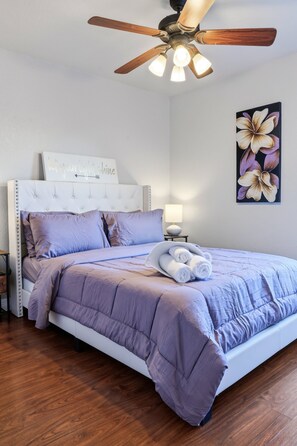 A bedroom with a tufted headboard, a bed with a purple comforter, a ceiling fan, and a wall-mounted flower painting. - Figaro - A Birdy Vacation Rental (San Antonio)