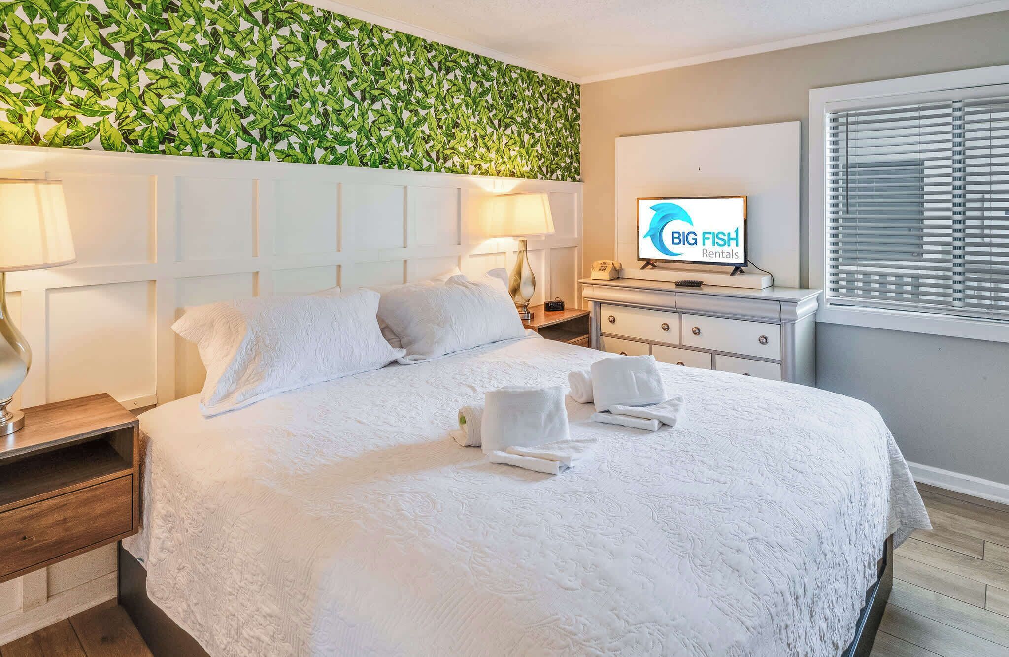 Una habitación de hotel con una cama grande, un televisor de pantalla plana, un teléfono montado en la pared y una ventana con persianas.