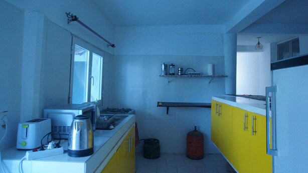 Una cocina con estufa a gas, hervidor, tostadora y pileta. Hay una ventana que proporciona luz natural y una alacena con utensilios de cocina.