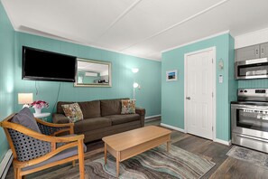 A living room with a sofa, a wooden chair, a coffee table, a flat-screen TV, a wall-mounted mirror, and a kitchen area with an oven. - Cozy Ocean Block Condo | Walk to Beach (Ocean City)