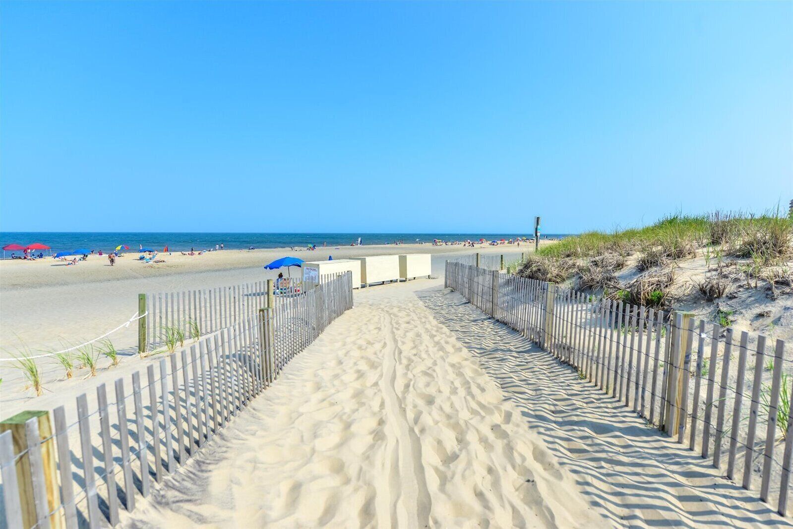 A sandy beach path leading to the ocean with a wooden fence and beach umbrellas.