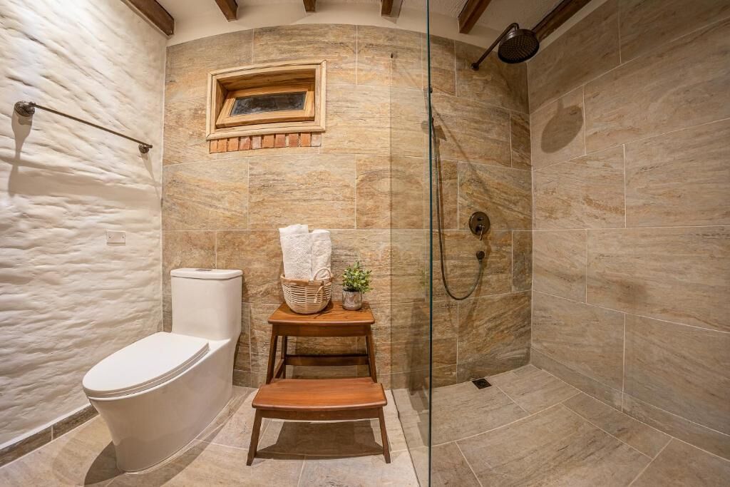 A bathroom with a toilet, a wooden stool, a small table with a basket and a plant, and a shower area with a glass door.