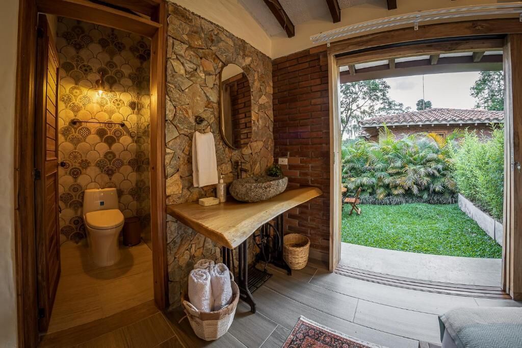 Un baño con pared de piedra, un banco de madera, un lavabo y vistas a un jardín.