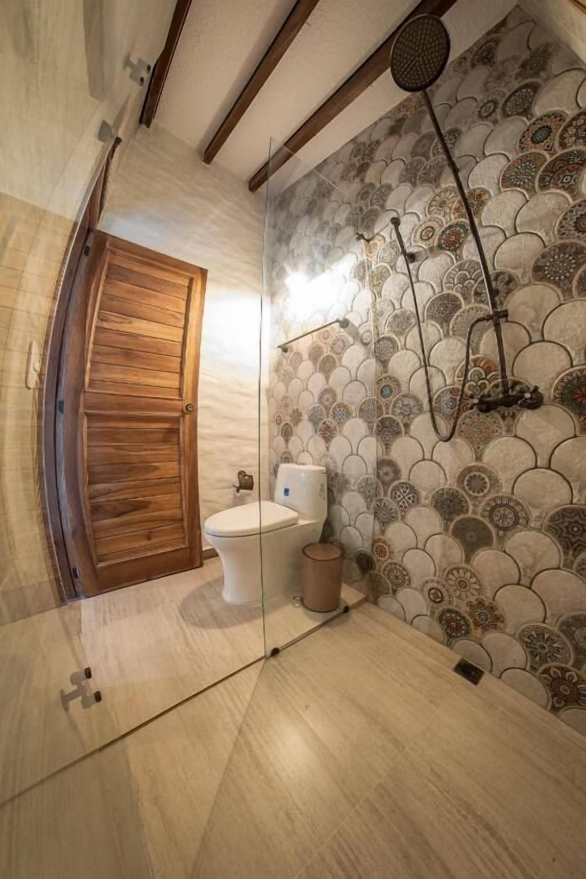 Un baño con puerta de madera, inodoro, zona de ducha con pared decorativa y una lámpara.