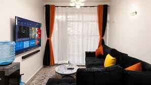 A modern living room with a flat-screen TV mounted on the wall, a black sofa with orange cushions, a round coffee table, and a large window with sheer curtains. - Luxury one bedroom-Roysambu near CBD (Nairobi)