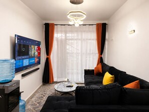 Ein modernes Wohnzimmer mit einem an der Wand befestigten Flachbildfernseher, einem schwarzen Sofa mit orangefarbenen Kissen, einem runden Couchtisch und einem großen Fenster mit transparenten Vorhängen.