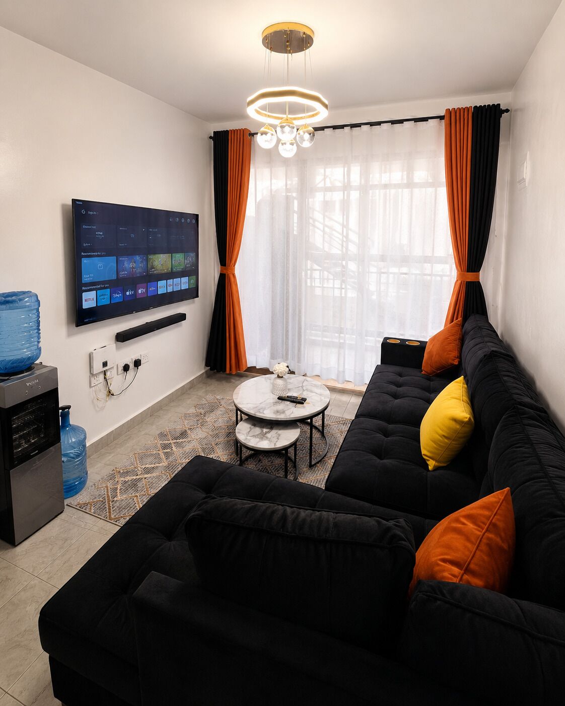 Ein modernes Wohnzimmer mit einer schwarzen Couch, einem Couchtisch, einem an der Wand befestigten Fernseher und einem großen Fenster mit Vorhängen.