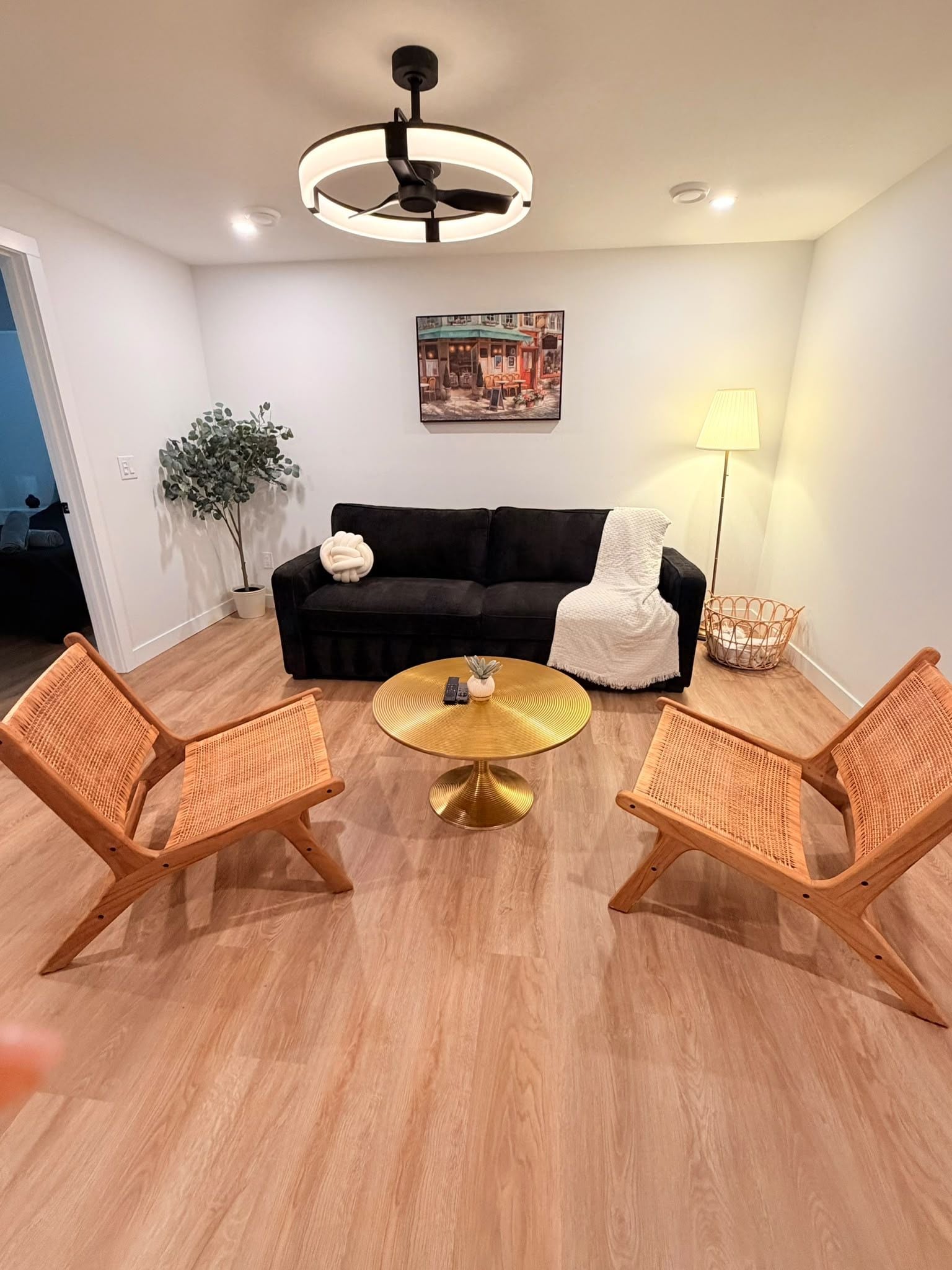 A modern living room with a black sofa, two wicker chairs, a round gold coffee table, a floor lamp, and a ceiling fan.