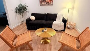 A modern living room with a black sofa, two wicker chairs, a round gold coffee table, a floor lamp, and a ceiling fan.