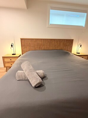 Een net opgemaakt bed met een grijze lakenset en twee opgerolde handdoeken, een houten hoofdbord, nachtkastjes met lampen en een raam met zonwering.