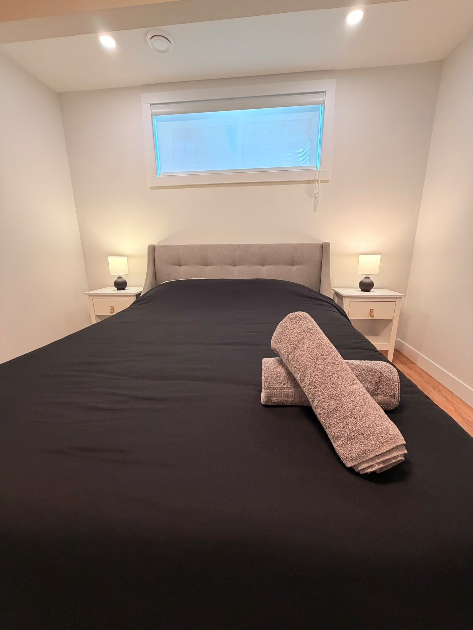 Ein Schlafzimmer mit einem großen Bett, zwei Nachttischen mit Lampen, einem Fenster mit Jalousien und einer Deckenleuchte.