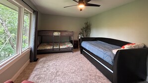 Quarto com beliches, ventilador de teto, janela com vista para árvores e uma planta.