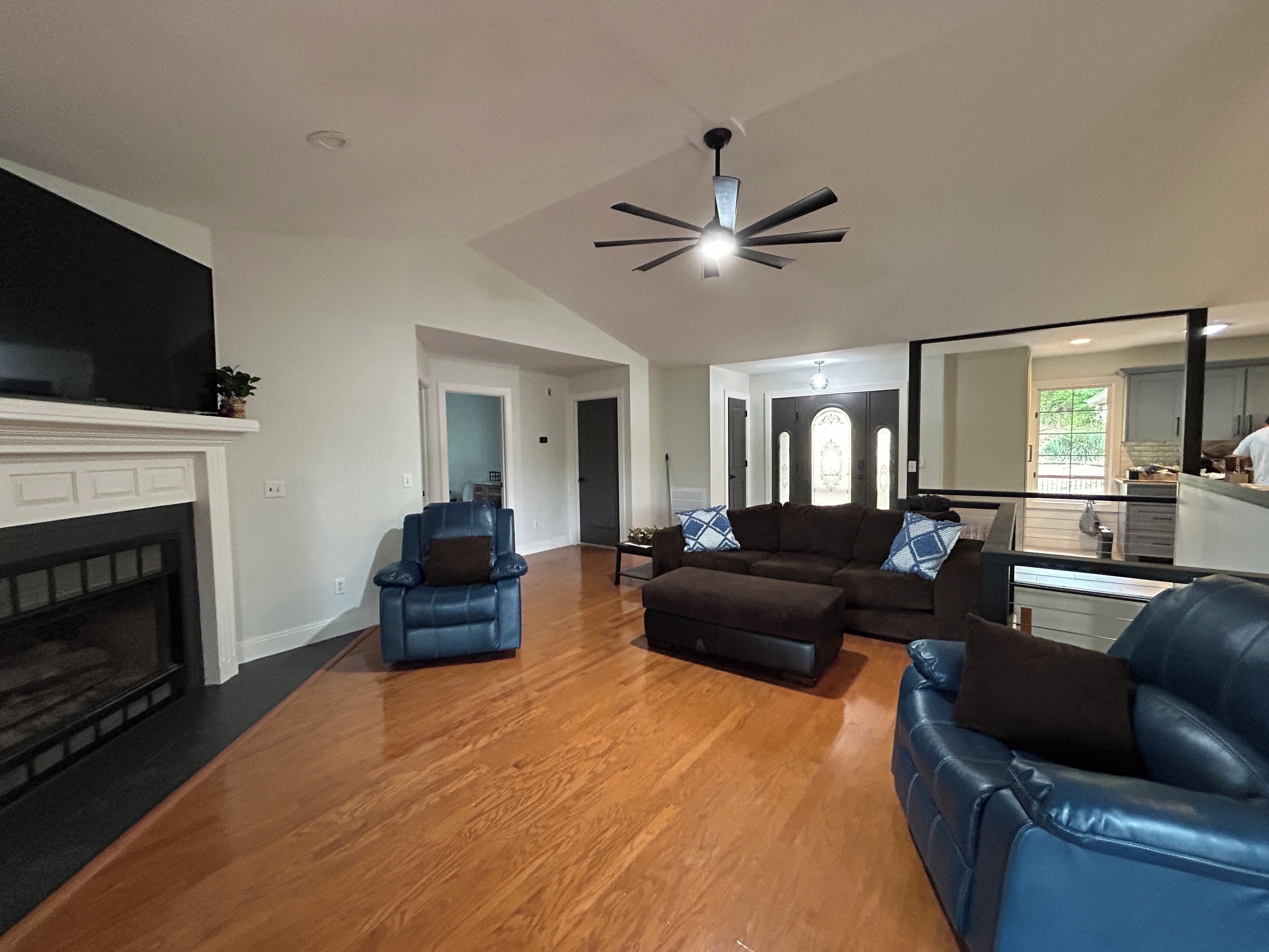A living room with a fireplace, a ceiling fan, and a view into the kitchen.