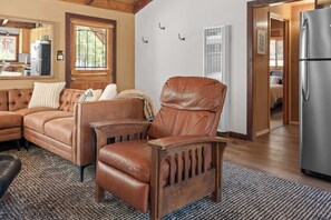 Ein Wohnzimmer mit einem Ledersessel, einer Couch und einem Couchtisch aus Holz. Im Hintergrund ist eine Küchenzeile erkennbar.