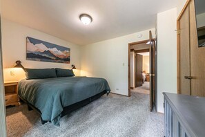 Ein Schlafzimmer mit einem Bett, einem Nachttisch, einer Wandleuchte, einer Tür zu einem anderen Raum und einem Bild an der Wand.