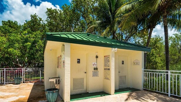Um pequeno edifício térreo com telhado verde e paredes brancas, com área de estar coberta, lixeira e um pequeno aparelho de ar condicionado.