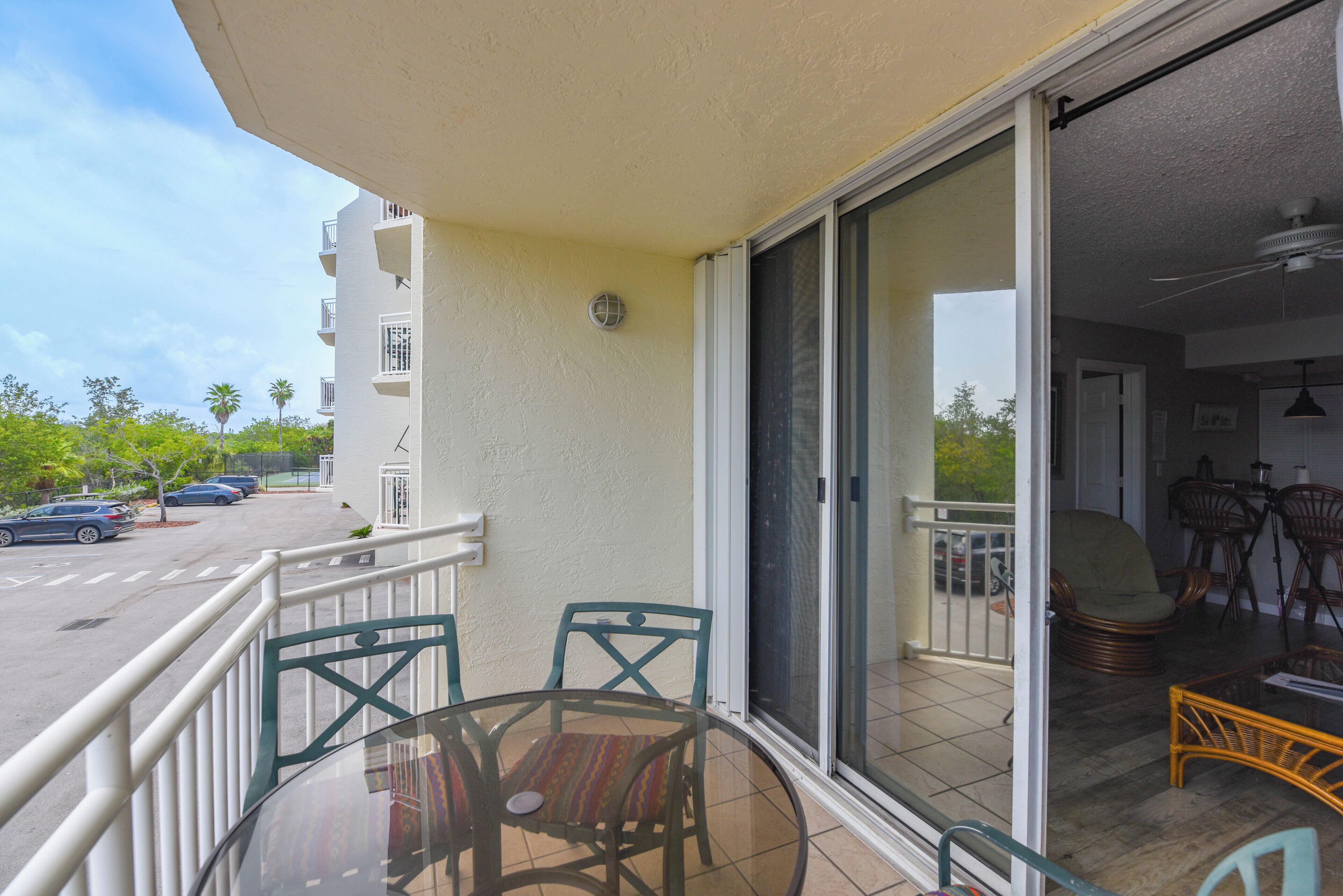 A balcony with a glass table and chairs, a sliding glass door leading to a patio area with outdoor furniture, and a parking lot with cars in the background.