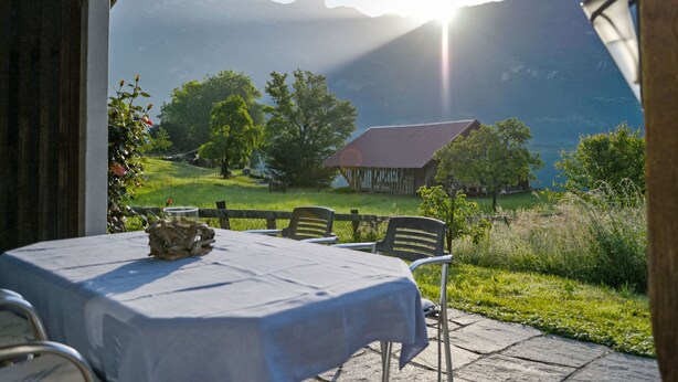Un área de comedor al aire libre con una mesa preparada para dos, con vistas a un paisaje montañoso y una cabaña de madera a lo lejos.
