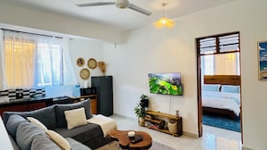 A modern living room with a sofa, coffee table, and a TV. There is a kitchen area with a refrigerator and a dining table. The room has a ceiling fan and a window with curtains. - Peaceful home at the heart of Kilifi town, with close proximity to the beach (Kilifi)