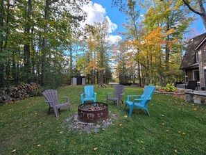 Ein Garten mit einem Feuerkorb, drei Adirondack-Stühlen und einem Holzstapel.