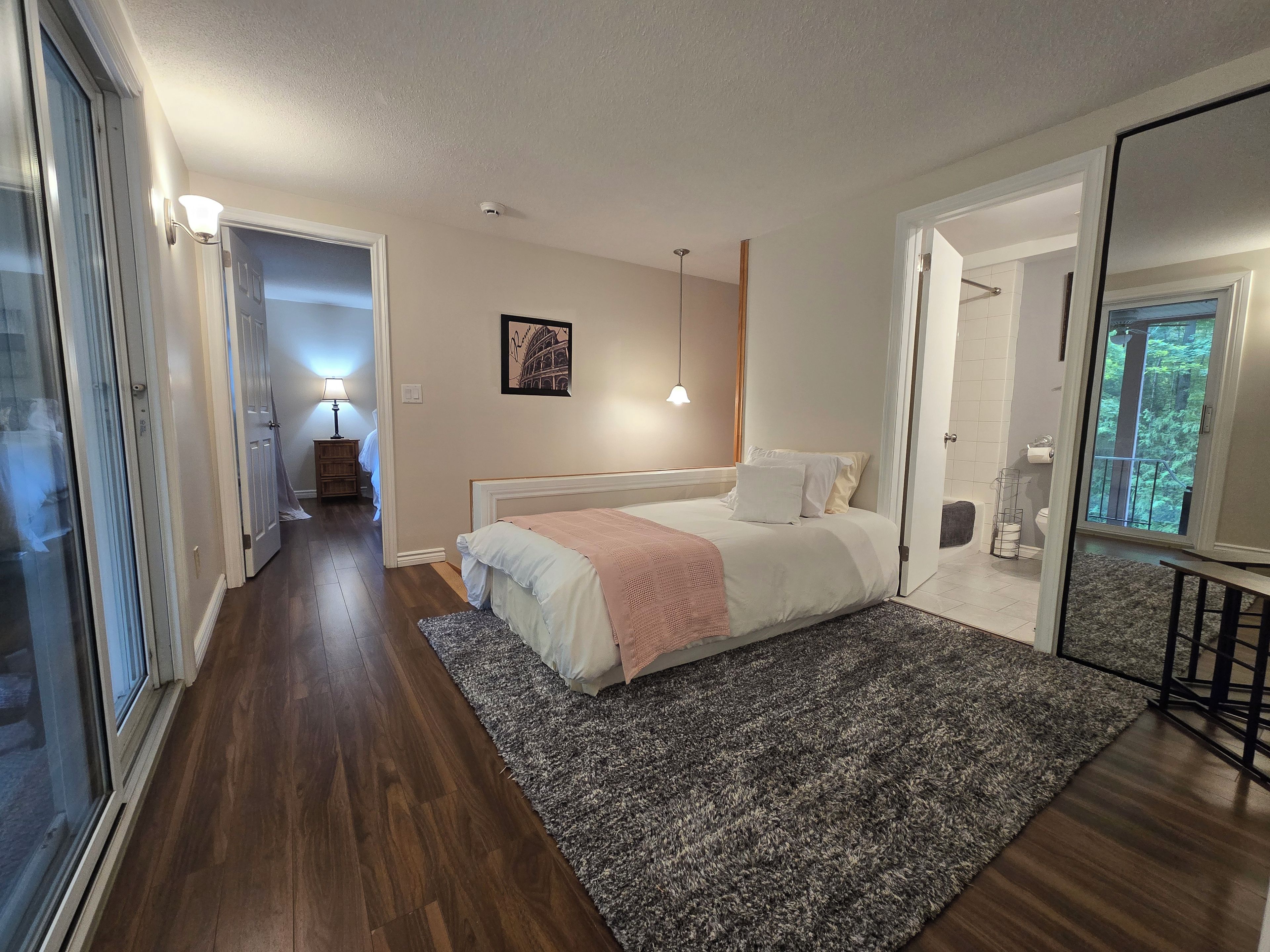 A bedroom with a bed, a nightstand, a lamp, a picture on the wall, and a view of the outdoors through a window.