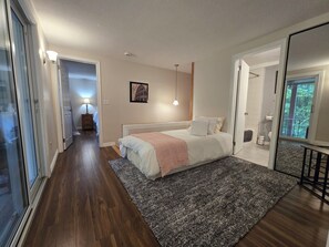 Un dormitorio con cama, mesita de noche, lámpara, un cuadro en la pared y vistas al exterior a través de una ventana.