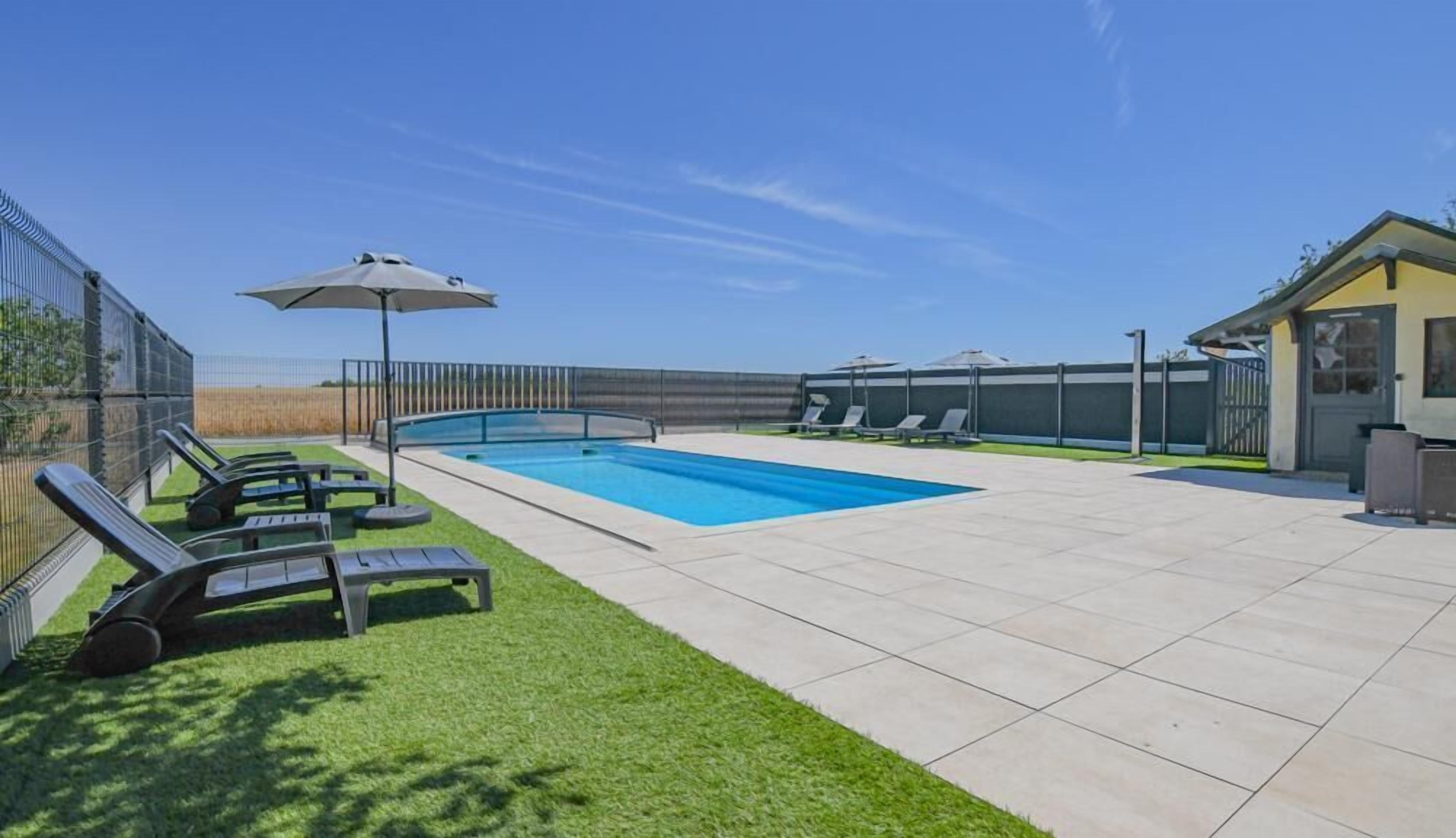A swimming pool with lounge chairs and umbrellas, surrounded by a fenced area and a building.