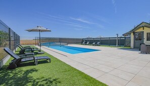 Una piscina con sillas de descanso y sombrillas, rodeada por una zona vallada y un edificio.