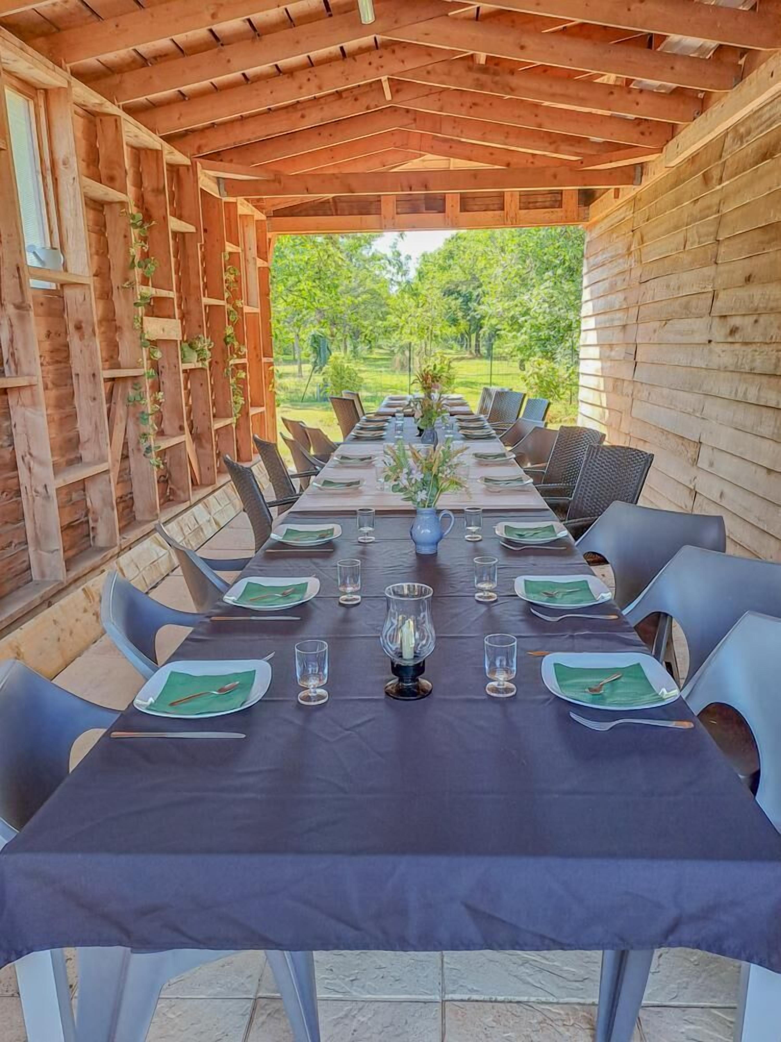 A covered dining area with a long table set for a meal, surrounded by chairs.