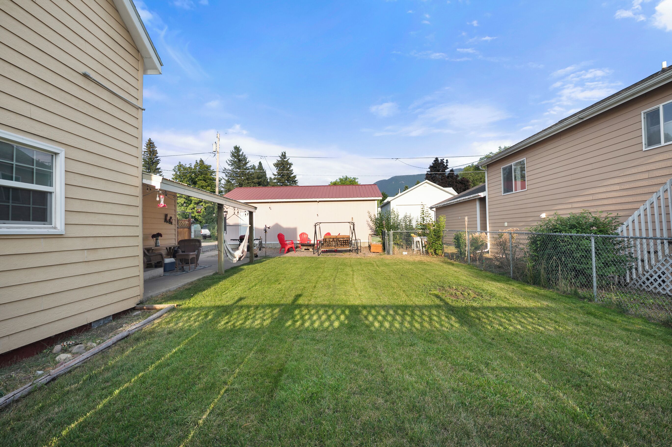 A residential backyard with a lawn, a patio with outdoor furniture, and a red-roofed building in the background.
